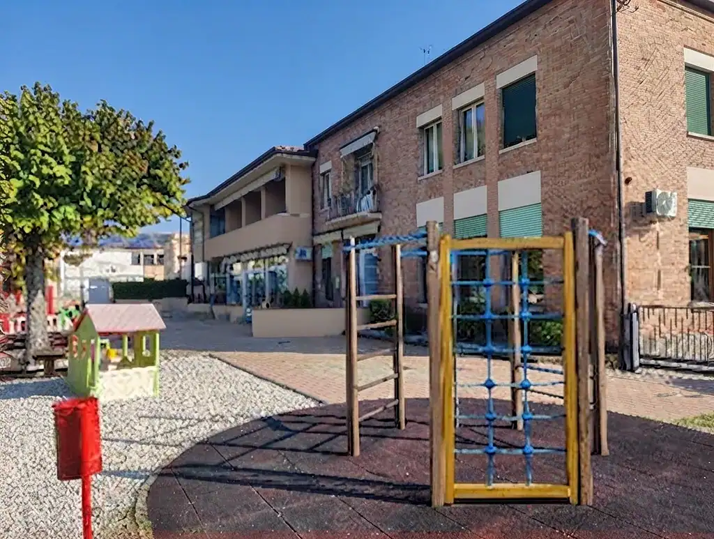 Spazio giochi all'aperto con attrezzature colorate e scuola in background.